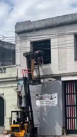 The video shows two men working on the upper floor of a two-story building. They are using a yellow forklift to reach the window, where one man is working. The other man is standing on the forklift, seemingly painting or working on the building's facade. The building is painted in a light color, with decorative vertical lines and several windows. Adjacent to the building is a gray structure with a sign and a multi-colored roll-up door. The street in front of the building has traffic, including black and white cars. The sky is partly cloudy.