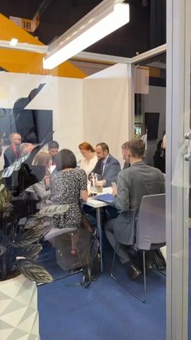 The video shows a group of people seated around a table in a well-lit room, likely an indoor event or exhibition, given the modern aesthetic and the presence of trade show booths in the background. The subjects are engaged in a discussion or meeting.

At the table, there are at least six individuals. In the foreground, a woman with dark hair and a patterned dress is seated, facing away from the camera. To her left, a woman with red hair pulled back is wearing a white blouse and appears to be listening. A man in a dark suit and tie, with a beard, is seated next to her, looking towards the left side of the frame. Further to the right, two other men are also present, one in a grey suit and the other in a darker suit, both looking down at papers or their phones.

On the table, there are water bottles, cups, papers, and a blue folder, suggesting a formal meeting or presentation. A plant is visible in the lower left corner, adding a touch of greenery to the scene. The overall atmosphere appears professional and focused. The lighting suggests it is daytime, and there are no indicators of weather conditions. The presence of trade show elements in the background hints at a public event or conference.

One of the visible elements in the background is a portion of a sign that reads "18". The overall setting is modern and clean, with white walls and bright overhead lighting.