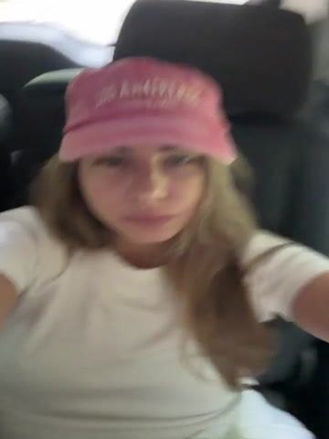 This media file captures a close-up selfie of a young woman sitting in the back seat of a vehicle. She is wearing a white t-shirt and a pink baseball cap that has the words "LOS ANGELES" embroidered on it in a lighter pink font. Her hair is long and light brown, and she is looking directly at the camera with a neutral expression, occasionally pursing her lips. The lighting suggests it could be daytime, possibly overcast given the soft light. The background is blurred, showing hints of dark upholstery from the car seat and a greenish blur outside the window, which might indicate trees or foliage, suggesting a road trip or travel through a semi-rural or natural environment. The woman's focus is entirely on the camera, implying she is taking the selfie for personal use or to share with someone. The overall mood is casual and relaxed. The visible text on the hat is the most prominent detail other than the subject herself.