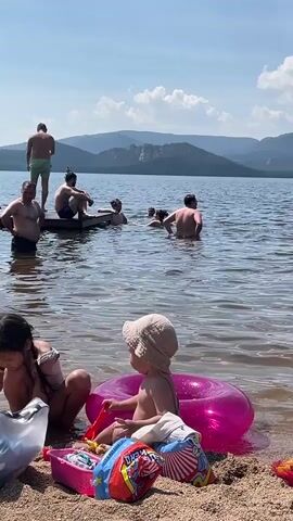 The video opens on a bright, sunny day at a lake. The foreground is dominated by a sandy beach where two young children are playing. A girl with dark hair is sitting down, looking at something in a bag. Next to her, a toddler wearing a light-colored hat sits beside a large, inflatable pink ring and a pink toy bucket containing various beach toys, including a red shovel. In the middle ground, several adults are in the water, swimming and wading. Some are standing on a wooden pier, looking out at the lake. In the background, a calm lake stretches towards distant, tree-covered mountains under a clear blue sky with scattered white clouds. The overall scene depicts a relaxed and pleasant day at the beach.