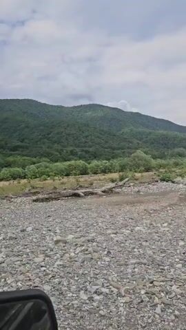 The video shows a view from inside a vehicle, looking out at a rocky riverbed and a forested mountain in the background. The sky is cloudy. The vehicle appears to be moving slowly, as indicated by the slight motion blur. The riverbed is covered with stones and gravel, and there are some fallen logs scattered around. The mountain is covered with lush green trees, and the slopes are gentle. The overall impression is one of a peaceful, natural landscape.