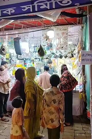 This video captures a bustling evening market or fair in Charfasson, Bangladesh. Numerous people, including men, women, and children, navigate the crowded aisles between market stalls. Many women are dressed in hijabs or other modest traditional clothing, and some men wear panjabis.

The stalls are covered by colorful overhead tarps and illuminated by strung lights, indicating the scene takes place at night or dusk. Various merchandise is displayed, with notable items including decorative hanging ornaments (possibly bird feeders) and colorful balloons, some shaped like footballs, dangling from the stall awnings.

People are actively walking through the market, browsing the offerings. One man is seen carrying a baby, and another person has a child on their shoulders, underscoring the family-friendly atmosphere.

Large banners featuring Bengali script are prominently displayed above the stalls. One banner clearly reads "...ফ্যাশন মাসব্যাপী..." (Fashion Month-long), followed by "...সাজিক সহযোগিতা..." (Social cooperation/assistance), and concluding with "...আয়োজনেঃ চরফ্যাশন" (Organized by: Charfasson), confirming the event's location and nature. Another banner briefly shows "মালিক সমিতি" (Owners Association).

A male voice speaking in Bengali can be heard, likely a vendor or announcer, calling out prices (e.g., 60 Taka, 195 Taka) and inviting people to "Come, come, see then..." The overall ambiance is lively and indicative of a popular local gathering or shopping event.