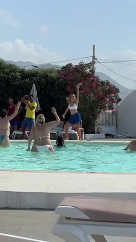 The media shows a lively scene in a swimming pool on a sunny day, likely in Capaci, Italy. The main focus is a group of people enjoying themselves. In the center of the pool, a young woman in athletic wear, a white sports bra and blue shorts, is energetically dancing on a raised platform, with one arm raised and the other extended. She appears to be leading an activity.

Around her, several other people are in the pool, participating or observing. To her left, a woman in a white bikini is also raising her arms, seemingly mirroring the dancer's enthusiasm. To the far left, a person in a green swimsuit is in the water, facing away from the camera. Further back, two men are visible; one in a yellow t-shirt and blue shorts is holding a camera or phone, appearing to record the event, while another man in a red shirt stands beside him. Another woman in a blue swimsuit is visible further back, near the edge of the pool.

The setting is an outdoor swimming pool with clear blue water and a white tiled edge. Lush green bushes and a tree with pink blossoms are visible behind the pool, with a hint of mountainous terrain in the background under a bright blue sky, suggesting a warm and pleasant climate. The overall atmosphere is one of fun and recreation, with smiles and energetic movements suggesting joy and a good time. There is no visible text in the media file.