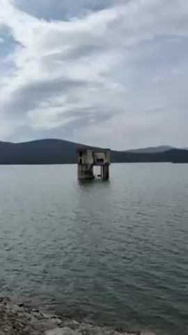The video shows a concrete structure standing in the middle of a lake. The structure looks like a small, two-story building, possibly a water pumping station or intake structure. It has visible windows and a stairway leading to an upper level. The structure appears weathered and old, with visible signs of decay and discoloration. The surrounding water is slightly choppy, and the sky overhead is overcast, suggesting a cloudy or possibly rainy day. In the background, there is a mountain range visible. There are ripples on the water, indicating slight wind movement. The camera zooms in on the structure during the video.
