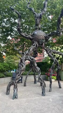 This video displays a kinetic sculpture in an outdoor setting. The sculpture, made of dark metal, resembles a cluster of intertwined branches supporting a large, textured sphere at its center. The sphere has a vaguely spherical or bulbous shape, with a patterned texture that suggests an organic or celestial body. The branches twist and turn, some extending upwards like arms, while others form multiple leg-like supports at the base. The sculpture stands on a light-colored gravel surface, with a paved pathway curving around it. In the background, lush green trees and shrubs provide a natural backdrop. Several large potted plants containing white flowers are visible, along with glimpses of red flowers in planters. A dark blue chair sits to the left of the sculpture, and further back, there are hints of buildings with reddish-brown brickwork and windows. The lighting suggests it might be dusk or a heavily overcast day, as the light is soft and diffused. The camera pans slowly, giving a full view of the sculpture and its surroundings, emphasizing the intricate details of the metalwork and the contrast between the artificial sculpture and the natural environment.