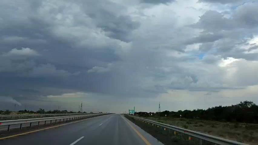 The video captures a view from a vehicle traveling on a wet highway under a stormy sky. Dark, heavy clouds dominate the upper portion of the frame, with some lighter patches and a hint of blue sky visible. A distant storm appears to be visible on the horizon, with a visible rainfall or dark cloud mass. The highway stretches ahead, appearing wet and reflective. Guardrails line both sides of the road, and distant power lines and vegetation are visible on the sides. Another white car is seen ahead, driving in the same direction, kicking up spray from the wet road. Road signs are visible in the distance. The overall impression is one of driving through potentially inclement weather.