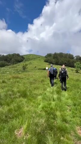 The video shows three individuals walking up a grassy hillside on a trail. The sky is blue with white, puffy clouds. The terrain is covered in lush green grass and scattered trees. The people are wearing backpacks and appear to be hiking. The camera is positioned behind them, following their movement up the slope. There is a signpost visible in the distance on the left side of the trail. The overall atmosphere is one of outdoor recreation and exploration.
