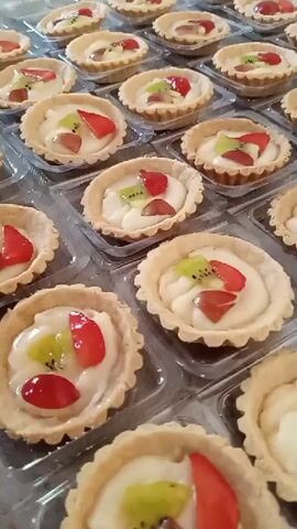 The video shows an overhead, vertical shot of a large quantity of fruit tarts arranged in clear plastic containers. The tarts are small and individually portioned, with a golden-brown, crimped crust filled with a white, creamy filling. Each tart is topped with a colorful assortment of fresh fruit, including sliced strawberries, kiwi, and red grapes. The tarts are neatly organized in rows and columns, filling the frame. The lighting is bright, highlighting the details of the tarts and their toppings. There are no people or animals visible in the video. The background is indistinct, suggesting the tarts are likely on a table or counter.