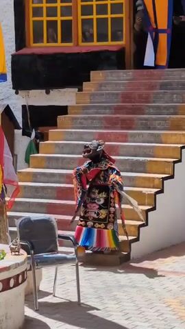 A traditional Tibetan masked dance is taking place. Several individuals, dressed in ornate and colorful robes, are present. One dancer in the foreground wears a vibrant, patterned costume and an elaborate mask, performing a ritualistic dance on a paved courtyard. Behind them, a set of stairs with red and yellow markings leads up to a building with yellow-framed windows. Another person in a red robe is partially visible on the stairs. Colorful prayer flags are strung from poles, adding to the festive atmosphere. A blue chair sits unoccupied in the foreground, to the left of the main dancer. The scene appears to be outdoors, in a courtyard likely associated with a monastery or temple.