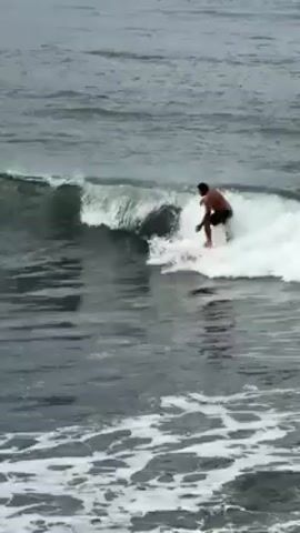 A man is surfing on a light-colored surfboard in a choppy, grey ocean. He is shirtless and wearing dark shorts. The waves are small and foamy, and he maneuvers the board smoothly across the water. Another surfer, wearing a pink top, is briefly visible paddling further out.