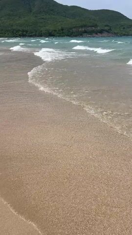 The video shows a beach with clear, shallow water lapping onto the sandy shore. Gentle waves are breaking in the distance. In the background, there are green, tree-covered hills or mountains. The sky appears partly cloudy. The camera is positioned low, close to the water's edge, and appears to be moving slightly, capturing the ebb and flow of the tide. There are no people or discernible objects on the visible portion of the beach or in the water. The scene conveys a sense of natural tranquility.