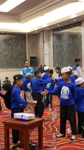 The video shows a group of children, mostly boys, wearing blue shirts and some wearing kufi hats, gathered in a room with red carpeting. They appear to be participating in an activity or game. Several adults are also present, some overseeing the children. In the foreground, a projector is set up on a wooden table. Some children are facing each other, extending their hands, possibly in a greeting or a game of hand-clapping. Others are standing around, watching or waiting their turn. The environment suggests an indoor event, possibly a gathering, workshop, or celebration.