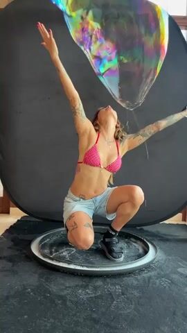A woman in a pink and white checkered bikini top and denim shorts is crouching on a circular platform that has white powder on it. She has tattoos on her arms and legs. Her arms are raised and spread wide, and she is looking up. In the background is a large black circular backdrop, and to the right of the backdrop is a window. She is holding a small black object in her right hand. Another person's hands are visible on the left side of the frame, holding a wire loop that is creating large soap bubbles. The woman is interacting with the bubbles as they float around her.