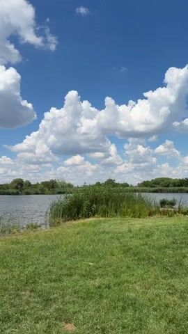 A vertical video pans across a scenic view of a body of water on a sunny day. The sky is a vibrant blue, dotted with fluffy white clouds. The foreground is a lush green grassy bank. The water is calm and reflects the sky. Along the edge of the water, there is dense green vegetation, including reeds. In the middle ground, a small fishing setup with rod holders is visible on the grass. Further to the left, a small, weathered boat rests partially submerged in the shallow water near the bank. The background is a line of trees and some distant buildings. The video captures a peaceful, natural setting.