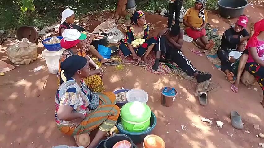 The video shows a group of at least nine people sitting on the ground in an outdoor setting. The ground appears to be mostly red soil. The people are a mix of men and women, all dressed in casual clothing. Many are sitting on mats or blankets. Several are engaged in what appears to be food preparation, with bowls and containers of food items scattered around them.  One man is lying down on a mat. Trees and vegetation are visible in the background. There are buckets, a small tub, and other miscellaneous items scattered around the group.  Someone is filming the scene, walking around the seated people. The lighting appears to be natural daylight. The atmosphere seems relaxed and communal.