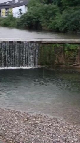 A drone flies over a shallow, rocky river. The drone moves from left to right, showing a concrete bridge spanning the river, then a stone wall with a drain pipe emptying water into the river, and finally the riverbed leading away from the drain pipe. The river is surrounded by trees and buildings.