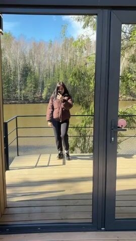 The video opens with a reflection of a woman in a large glass door. She is wearing a brown puffer jacket, black trousers, and black shoes, holding a mobile phone, likely taking a selfie or recording. The reflection reveals a wooden deck, a serene body of water, and a dense forest under a clear blue sky. A black door frame with a handle and a small pink circular sticker is visible on the glass.

The camera then pans right, showcasing the exterior of a modern cabin. It features large floor-to-ceiling glass doors and windows with dark frames, opening onto a spacious wooden deck. The deck is furnished with a small black round table and two matching chairs, offering an outdoor seating area. The cabin itself has dark gray siding and wooden accents.

Further right, the view expands to include the natural surroundings. Another similar modern cabin with a dark exterior is visible in the distance, nestled among the trees. The setting is a forest located beside a body of water, likely a lake or river, characteristic of Mishkino, Russia. Tall pine and fir trees dominate the landscape, their branches reaching towards the sky. The camera captures the tranquil waterscape and the lush greenery from the perspective of the cabin's deck. Sunlight illuminates the scene, casting shadows on the wooden planks.

Faint, unreadable text is briefly visible on the glass door at the beginning of the video. The overall impression is a peaceful, modern retreat integrated into a natural, forested waterside environment.