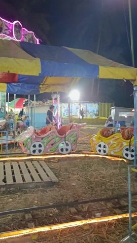 The video shows a carousel ride at night, likely at a local fair or carnival in Sambirata, Indonesia. The ride features colorful, cartoon-like vehicles, including cars and possibly other whimsical shapes, that spin around a central pole. The vehicles are painted in bright colors like yellow, green, and pink, and have large, circular wheels. Some of the vehicles are decorated with painted images of animals and plants.

Several people are visible on the ride. In the foreground, a young woman with her hair in a ponytail is smiling as she looks down. Further back, a man is seated in one of the cars, looking towards the camera with a relaxed expression. Other individuals can be seen in the background, either on the ride or observing it.

The setting appears to be outdoors, with dry grass and dirt on the ground. The background includes a large, striped tarp, likely part of a tent or stall, and some brightly lit structures, including what appears to be a string of yellow lights running along the right side of the frame. A tall, decorative structure with pink neon lights is visible on the left side of the frame. The lighting suggests it is nighttime, and the air seems still, with no indication of wind. The overall atmosphere seems festive and relaxed, characteristic of a local community gathering.

Visible text includes "Coca-Cola" on a red crate in the foreground. There are also some indistinct labels or signage on the ride structures and vehicles, but they are not legible.