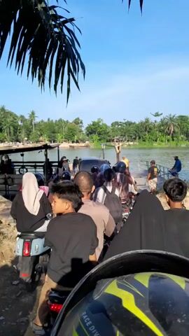 The video shows a group of people on motorcycles waiting to board a small ferry. The ferry is already partially loaded with people and vehicles. The scene is set on a river or lake, with green vegetation visible on the opposite bank. The sky is clear and blue. The camera appears to be mounted on a motorcycle, capturing the view from the perspective of someone waiting in line.