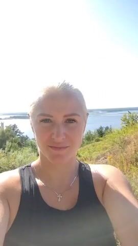 A woman with blonde hair pulled back, wearing a black tank top and a silver necklace, smiles at the camera. She is outdoors with a body of water and trees in the background under a bright, clear sky.