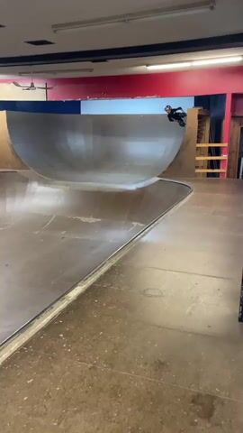 A person on a skateboard is seen skating inside a skatepark. The skatepark has a bowl-shaped ramp and wooden walls. The skateboarder is wearing a helmet, knee pads, and elbow pads. The skateboarder is performing a maneuver on the ramp. The scene is lit by overhead lights. The walls of the skatepark are painted red and blue.