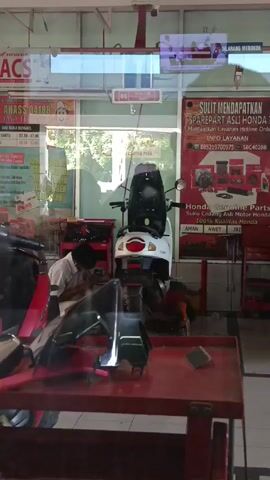 Here is a concise description of the video:

The video shows a view into a Honda motorcycle repair shop. A mechanic is working on a red motorcycle in the background. A white scooter is parked in the center of the frame. The shop has a red tool cart and a red table with motorcycle parts on it. There are signs on the windows with Honda logos and information about the shop. The overall impression is of a busy, well-equipped motorcycle repair shop.