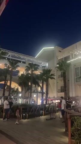 The video shows an outdoor area at dusk with palm trees and a large building with multiple levels. Several people are walking around. In the foreground, two people are walking on a wooden boardwalk. One person is carrying a bag. Further back, several other individuals are visible, including a woman and a child. Some people are standing and talking. The building is illuminated with string lights. The sky is dark blue, indicating it is nighttime or late evening.