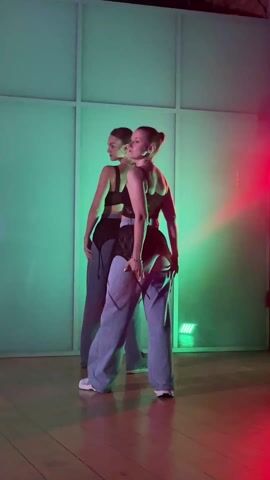 Two fair-skinned women are dancing in front of a geometric white paneled wall lit by green and red lights. They both wear black tops and baggy light blue pants. One dancer has her hair in a ponytail, while the other dancer's hair is down. Their dance includes arm movements and head turns.