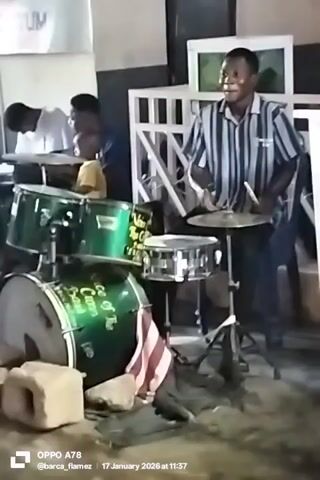 A man is playing the drums in a room. He is wearing a striped shirt and jeans. The drums are green, and there is a bass drum with text on it. There are other people in the room, including one in a white shirt who is writing something, and a child in a yellow shirt. The room is filled with furniture and other items. The video also has text at the bottom, including the brand of the phone that recorded the video and the date.