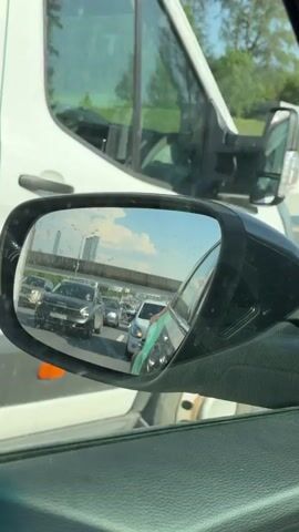 The video shows a car’s side mirror reflecting a highway traffic jam. Several cars are visible in the mirror, with a tall building in the background. The sky is blue with some clouds. The view is from inside a car, showing the driver’s side mirror and the edge of the car’s window. The traffic appears slow-moving or stopped.