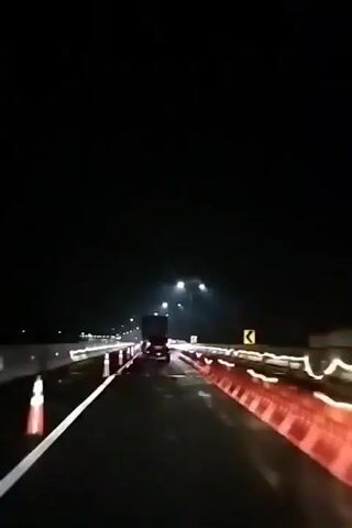 The video shows a nighttime view from inside a moving vehicle on a highway. The road appears wet, possibly from rain, and the dark sky is illuminated by streetlights and the headlights of other vehicles. In the distance, a truck is visible, with its brake lights illuminated. Orange traffic barriers and white traffic cones line the right side of the road, suggesting construction or a lane closure. Yellow caution signs with arrows indicating a curve are also visible. The vehicle's headlights are on, and the scene is blurry due to motion.