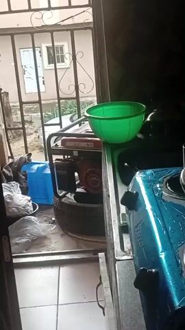 The media file shows an interior view from a doorway, looking outwards towards a barred window. Inside, a stovetop is visible, covered in blue protective plastic with black knobs. To the left, a green bowl sits on top of the stovetop. Further to the left, a red and silver generator is placed on a tire. Adjacent to the generator is a large blue water container. Several plastic bags filled with items are also on the floor near the generator.

Outside the barred window, a building with a light-colored facade and a white-paned window is visible. A car is parked in the area outside. The ground appears to be unpaved.

The scene suggests a functional, possibly outdoor or semi-outdoor kitchen area, common in some residential settings in Asubi, Nigeria. The presence of a generator and a water container implies a need for backup power and water supply, which can be typical in areas experiencing inconsistent utility services. The time of day is indeterminate, but the lighting suggests it is daytime. The overall mood is utilitarian and perhaps a bit cluttered, focusing on essential household items. No people are visible in the frame.
