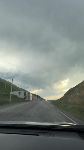 A car travels down a road on a cloudy day, passing a building on the left. Another car approaches and passes. Raindrops are visible on the windshield. Hills are on either side of the road.