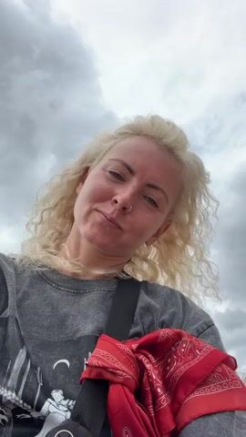 The video displays a close-up, slightly tilted selfie of a woman with blonde, curly hair, and a nose ring. She is wearing a grey t-shirt with a graphic print and has a red bandana draped over her shoulder. The background consists of a cloudy sky. In the first frame, she looks at the camera with a neutral expression. In the second frame, she sticks out her tongue playfully. In the third and fourth frames, she smiles broadly at the camera. The video is brief, with each frame lasting approximately one second.