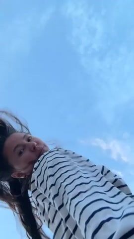 A low-angle, full shot shows a young woman with long dark hair wearing a black and white striped long-sleeved shirt and light-colored pants against a blue sky with some clouds. Her hair is blowing to her left. She is looking up and to her right. The woman extends her right hand forward towards the camera. She then raises her right arm above her head and her hair flies around her face. She then moves her arms widely as if dancing or exercising. The video is blurry and shaky in parts.