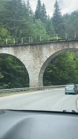 The video shows a view from inside a vehicle as it drives on a paved road. The road curves to the right under a large stone viaduct with two arches. The viaduct is made of rough-hewn stone blocks. Beyond the viaduct, a dense forest of green trees covers a hillside. Another vehicle, a light green hatchback, is visible on the road ahead, also driving under the viaduct. The sky appears overcast. The camera is positioned low, suggesting it is mounted on the dashboard or a similar surface inside the car. The lighting is diffused, consistent with an overcast day.