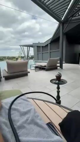 The video shows a first-person view of someone relaxing on a lounge chair by a rooftop pool. The person is lying down with their legs extended, and a grey towel is draped over their body. A hookah with a black hose sits on a small wooden table next to the lounge chair. Beyond the pool are several other lounge chairs, a modern building with a pergola, trees, and a lake under a cloudy sky. There is light music playing in the background.