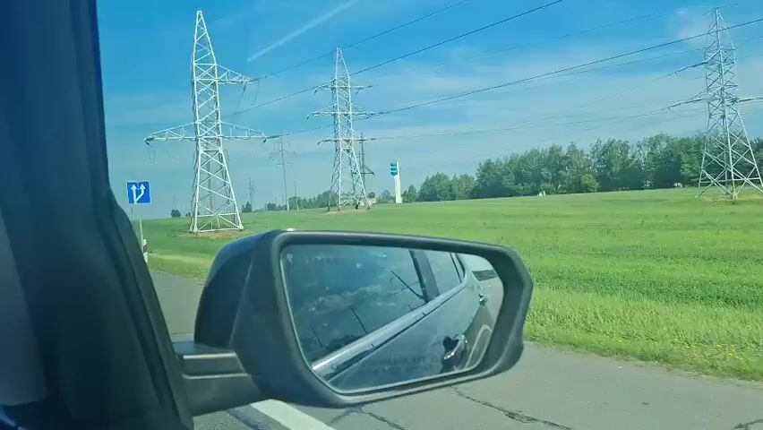 The video shows a view from inside a car, looking out the passenger side window and into the side mirror. The car appears to be on a paved road with a white line on the right edge. To the left of the road, there is a large, flat, green field. Several tall, white electrical pylons with power lines strung between them are visible in the distance, stretching across the landscape. A blue road sign with white arrows indicating a fork in the road is visible near the pylons. The sky is bright blue with a few faint white contrails. The side mirror reflects the car's interior and the side of the car itself, as well as the passing scenery. The overall impression is of driving through a rural area on a sunny day.
