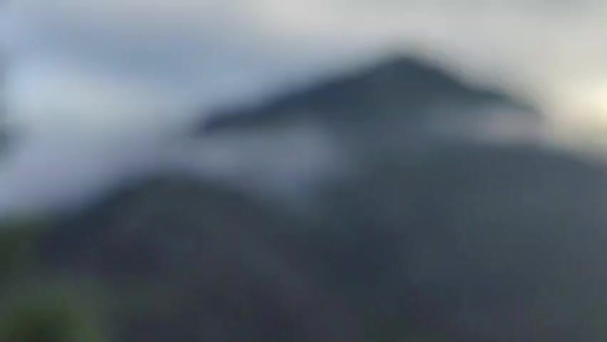 This is a medium shot of a misty mountain. The mountain is covered in green grass and has a line of trees along the top ridge. The fog is rolling down the side of the mountain in wisps. There are also power lines in the foreground of the shot. The sky is overcast and gray. The overall mood of the video is calm and serene.