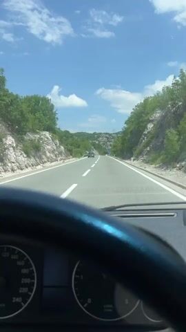 A first-person perspective video shows someone driving a Mercedes-Benz on a sunny day. The dashboard and steering wheel are clearly visible, as well as the speedometer and RPM gauge. The speedometer indicates the car is moving between 80 and 100 kilometers per hour. The RPM gauge is slightly above zero. Outside the car, the road stretches ahead, bordered by rocky hills and green trees under a blue sky with scattered clouds. The driver appears to be maintaining a steady pace on what looks like a highway. Music is playing in the background.