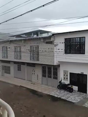 The video shows a street view on a rainy day, captured from a balcony. The street is wet, with water running down from building roofs and overflowing gutters. Two-story buildings line the street, with varying facades, some appearing newly constructed or renovated. A black motorcycle is parked in front of one of the buildings. The sky is overcast, and utility wires are visible overhead. Mountains are vaguely visible in the background, obscured by the heavy rain and mist. The balcony railing is visible in the foreground, partially obscuring the view.
