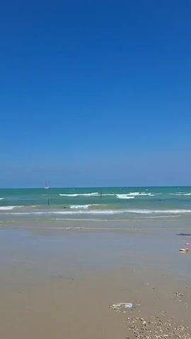 The video shows a wide shot of a beach with a bright blue sky overhead. Gentle waves lap onto a sandy shore. There are several pieces of debris scattered on the sand, including plastic items and what appears to be some seaweed or organic matter. Further down the beach, there are several boats moored on the shore. In the distance, a small cluster of buildings is visible, backed by a line of trees. The overall impression is of a tranquil, sun-drenched coastal scene, albeit with some evidence of human impact in the form of litter.