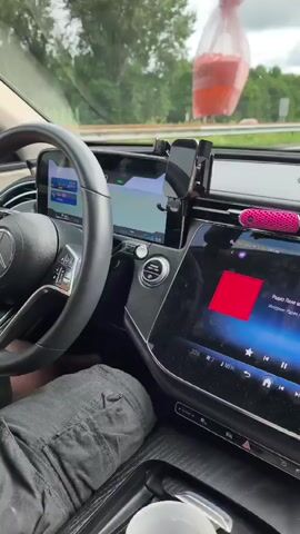 The video shows the interior of a car driving on a highway. The driver's side of the car is visible, with the steering wheel and dashboard in focus. The car is equipped with a large touchscreen display, a phone mount, and a pink air freshener. A small pink teddy bear is hanging from the rearview mirror. The road ahead is visible through the windshield, with trees and a cloudy sky in the background. The car appears to be in motion, as indicated by the blur of the scenery outside.