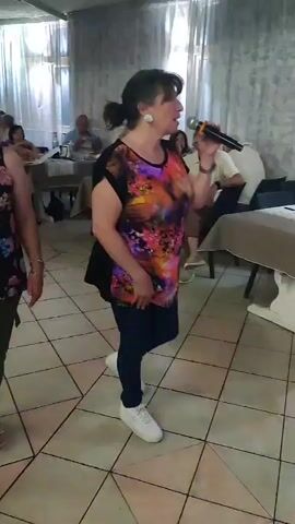 The video shows a woman singing into a microphone at an indoor event. She is wearing a colorful, patterned t-shirt, dark pants, and white sneakers. She has her hair pulled back in a ponytail and is wearing large earrings. Other people are visible in the background, seated at tables and observing the performance. The setting appears to be a room with tiled floors and draped curtains. The lighting suggests it is daytime, possibly indoors for a gathering or celebration. The woman seems engaged and expressive as she sings.