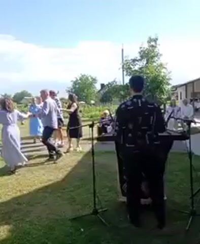 The video shows an outdoor gathering with people dancing in a circle. In the foreground, a person is standing behind what appears to be a keyboard or music setup, facing away from the camera. The dancers are a mix of adults and children, holding hands as they move. The environment is grassy with trees and buildings visible in the background under a bright blue sky with some clouds. The overall atmosphere appears festive.