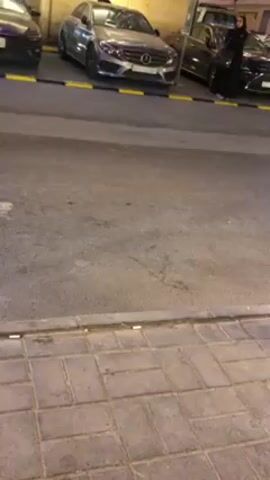 The video shows a view from a low angle of a parking area at night. There are three parked cars visible, a dark one on the left, a silver Mercedes in the center, and a dark SUV on the right. A woman dressed in a black garment is standing next to the SUV. The ground in the foreground is made of grey, rectangular paving stones and the parking area is marked with yellow lines. The general lighting is dim.