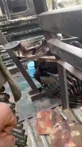 The interior of a vehicle is shown, possibly a military vehicle. Ammunition rounds are visible in a belt, likely for a machine gun. Metal frames are constructed within the vehicle, with what appears to be a seat and various tools. A person is partially visible inside the vehicle, dressed in dark clothing, next to a blue bucket. The vehicle's interior is cluttered with various items.