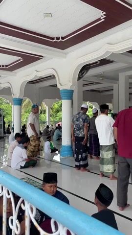 The video shows a group of Muslim men and boys in a mosque performing the prayer. The men are wearing traditional Islamic attire, including sarongs and prayer caps. The mosque appears to be a spacious, open-air structure with white pillars and a tiled floor. Some individuals are in the process of bowing, while others are sitting or standing. The atmosphere seems peaceful and devout.