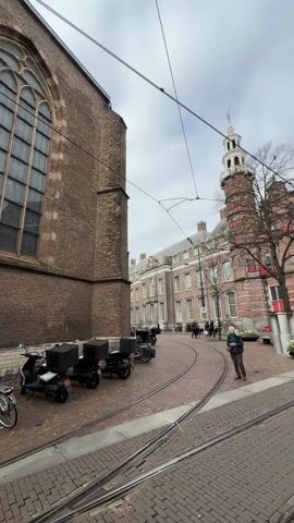 The video shows a street scene in The Hague, Netherlands, on an overcast day. The ground is paved with red bricks and has tram tracks running along it. A pedestrian with grey hair and a green scarf is walking towards the camera. Several black motor scooters with delivery boxes attached are parked along the side of the street. In the background is a large brick building with a tall, white-topped tower. A section of the building is decorated with red flags. Overhead are tram power lines. A person on a scooter passes by on the right.