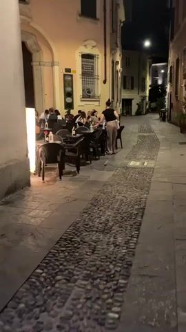 The video captures an outdoor dining scene at night. Several tables and chairs are set up on a cobblestone street. People are seated at the tables, appearing to be dining or socializing. One person, dressed in light-colored pants and a dark top, stands and appears to be serving or interacting with those at the tables. The area is illuminated by ambient lighting from the buildings and possibly streetlights, creating a warm and inviting atmosphere. The architecture suggests an urban European setting.